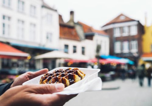 Tourist holds in hand popular street food - Belgium tasty waffle with chocolate sauce on the background of city tourist streets of Bruges, Belgium, Europe. Traditional Belgian dessert, pastry.